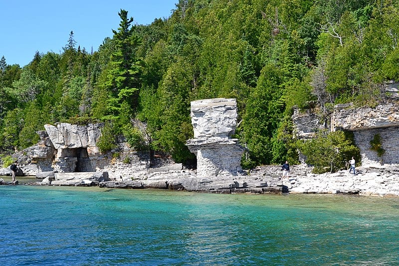 A coastal scene with bright blue water, granite cliffs edged with dense coniferous forests, and blue sky above.