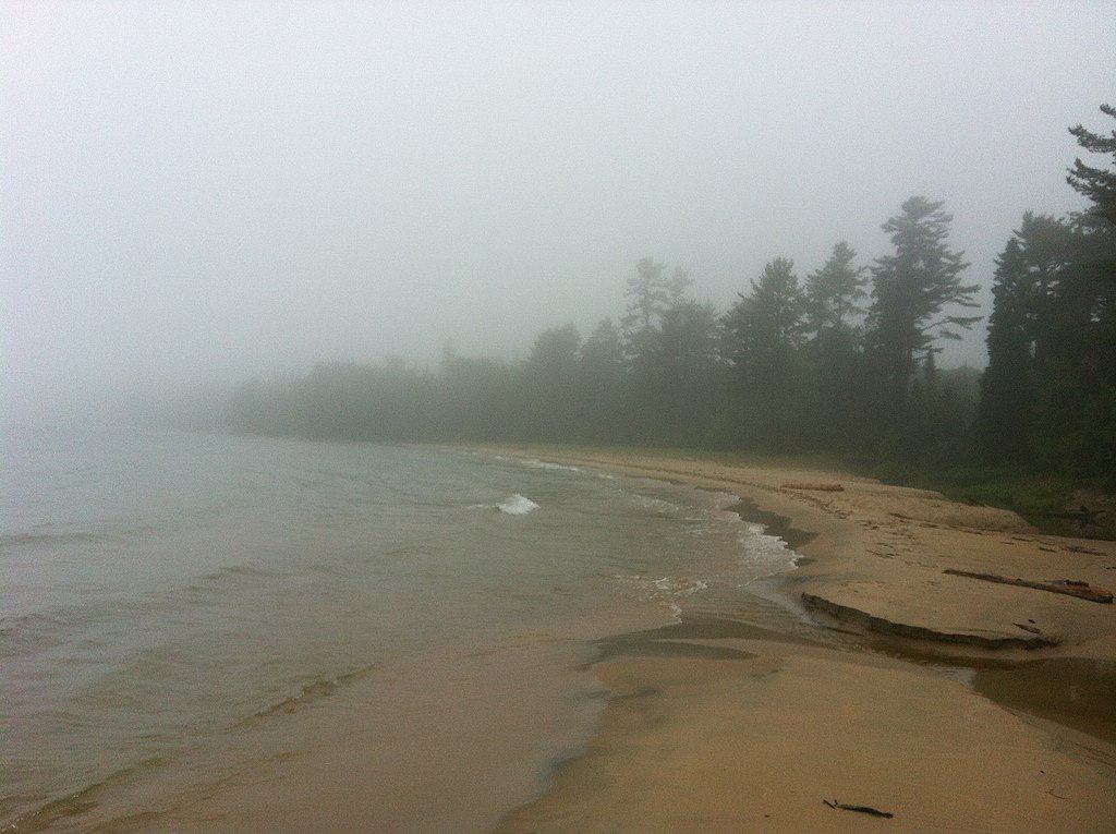 A foggy coastline with dark sand, tall pines, and fog rolling in off the water. (Katherine Cove, Lake Superior)