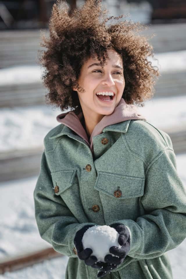 A woman in a green coat is smiling while holding a snowball.