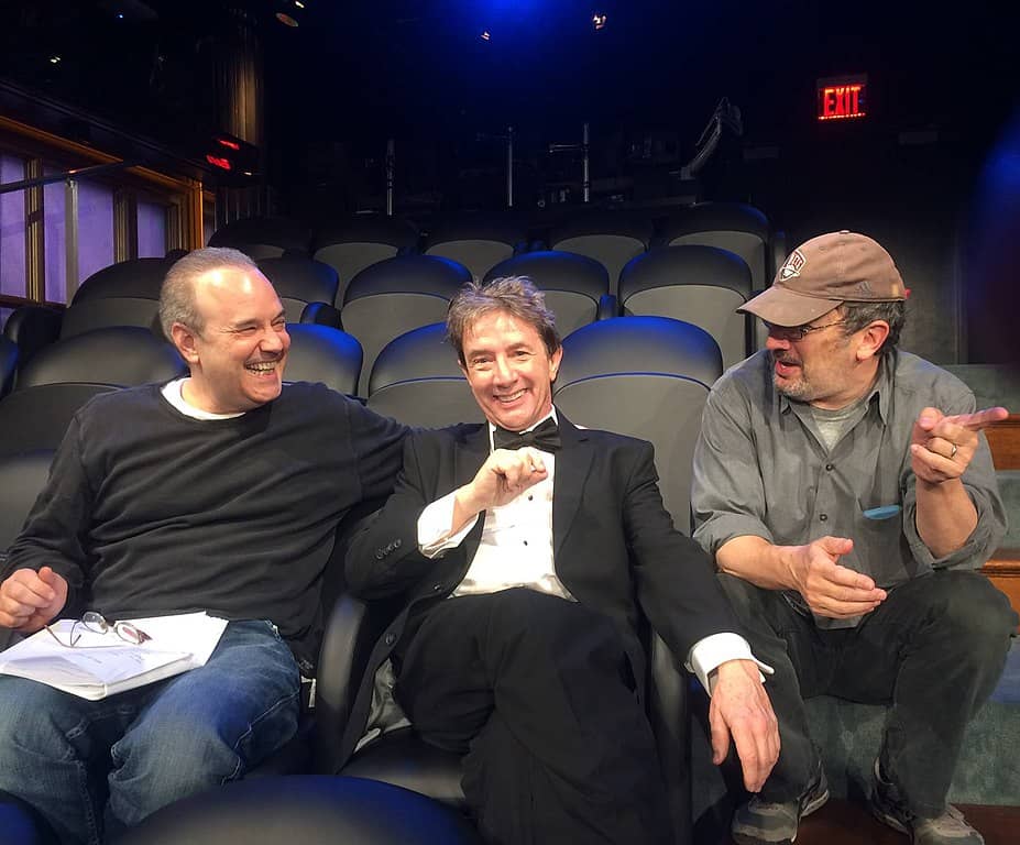 Three men sit on theatre seats, laughing. One of them points offscreen.