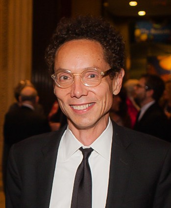 A man wearing a dark suit smiles and frameless glasses smiles at the camera