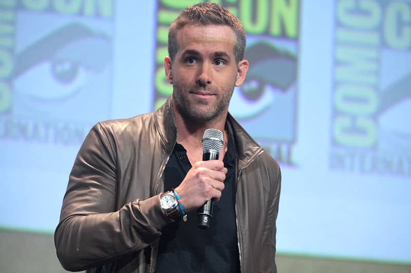 A blond man holds a microphone with an amused look, in front of a greyscale Comic-Con backdrop
