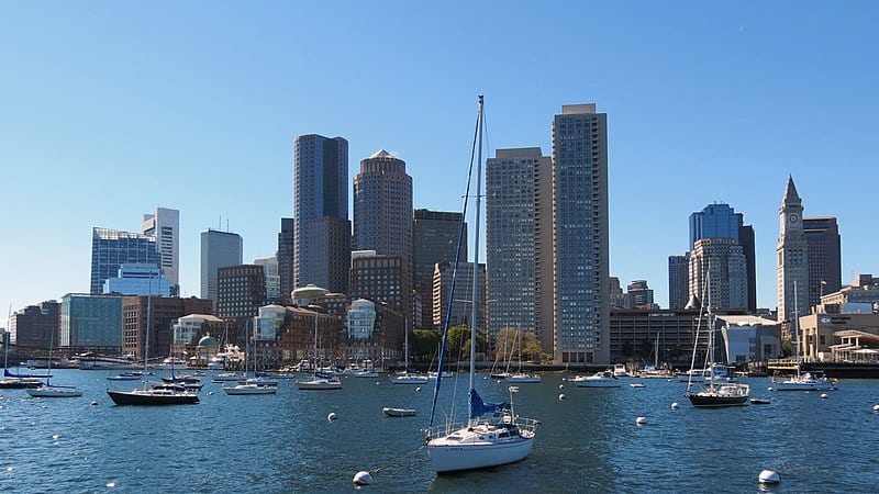 A harbour view of Boston (a large city with port buildings, skyscrapers, sailboats in the harbour) under a bright blue sky.