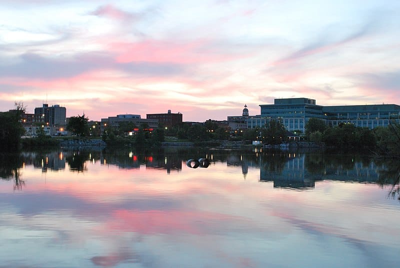 A view of Peterborough, Ontario at sunset (midsize city with trees and towers); the sky at sunset is mirrored in the Otonabee River.