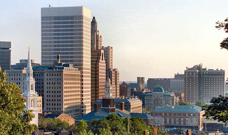 A skyline view of Providence: skyscrapers, church towers, brick mid-height buildings, framed gently by deciduous trees and a sky in early twilight.