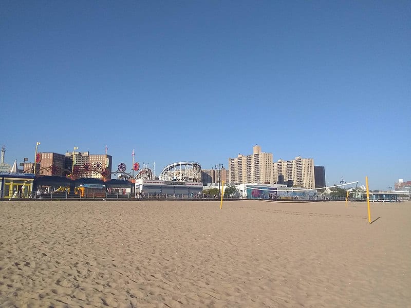 Daytime scene of a wide golden sand beach; an amusement park is beyond it with Ferris wheels and roller coasters, with apartment buildings behind it. (Coney Island, Brooklyn, NY)