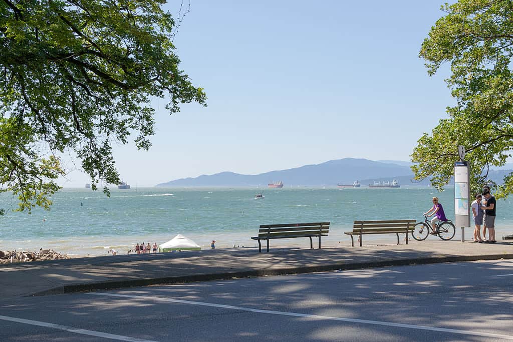 Landscape photo of English Bay (North Shore mountains in the distance, green-blue water, golden sand beach, with people looking at a signpost and two empty park benches in the foreground.) Vancouver, Canada.