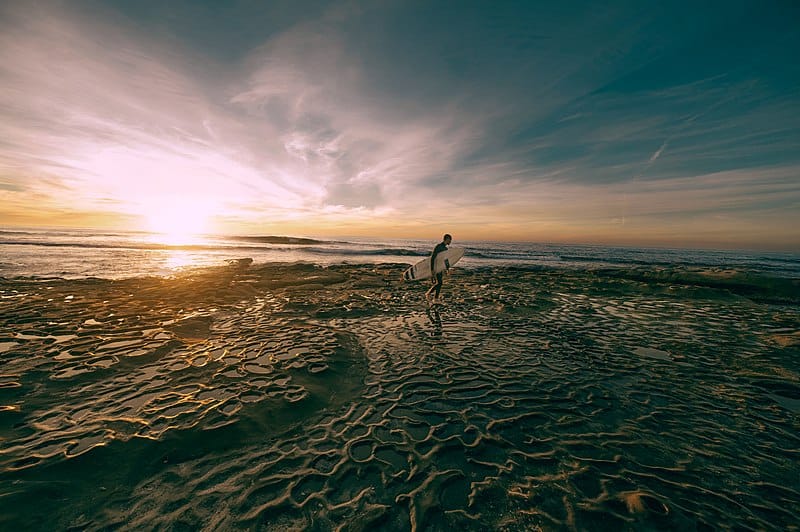 Seaside photograph; a receding tide washes over rippled sand/rock, with a surfer standing with his board on the horizon. (San Diego, CA)