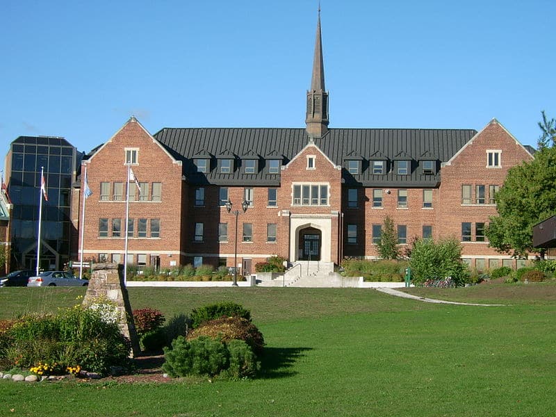 A three-storey red brick school building with a black roof and single spire on a green lawn, framed by trees. (Algoma University)