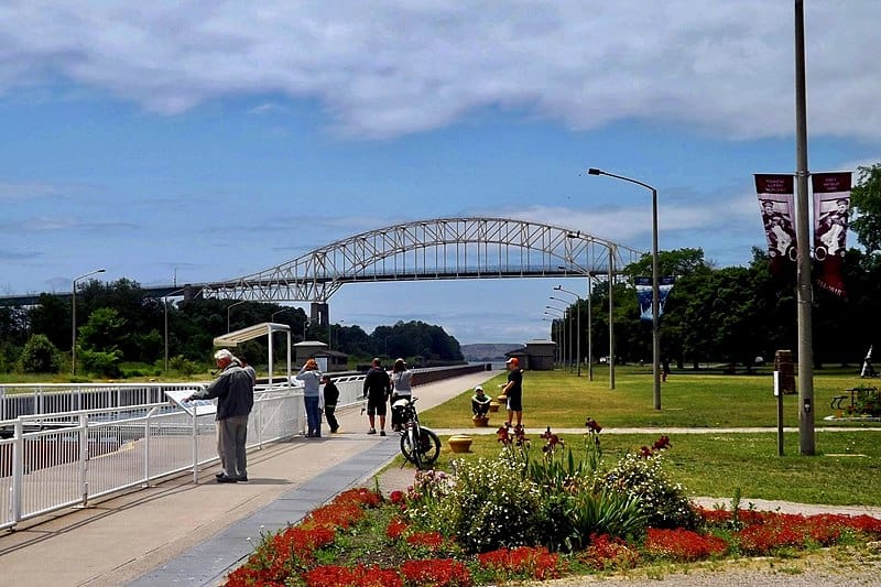 People walk along a riverbank and urban green space; at the horizon, a white metal bridge spans the river.