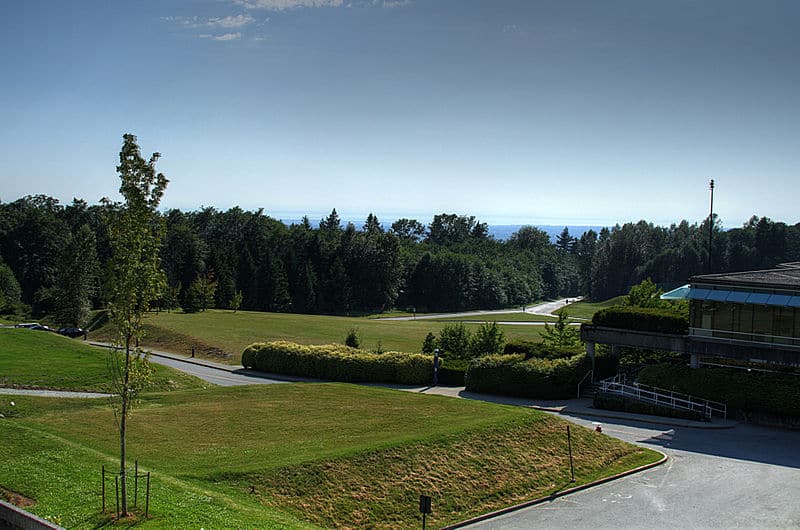 Rolling green hills with a pine forest on the horizon. In the foreground are some Simon Fraser University buildings.