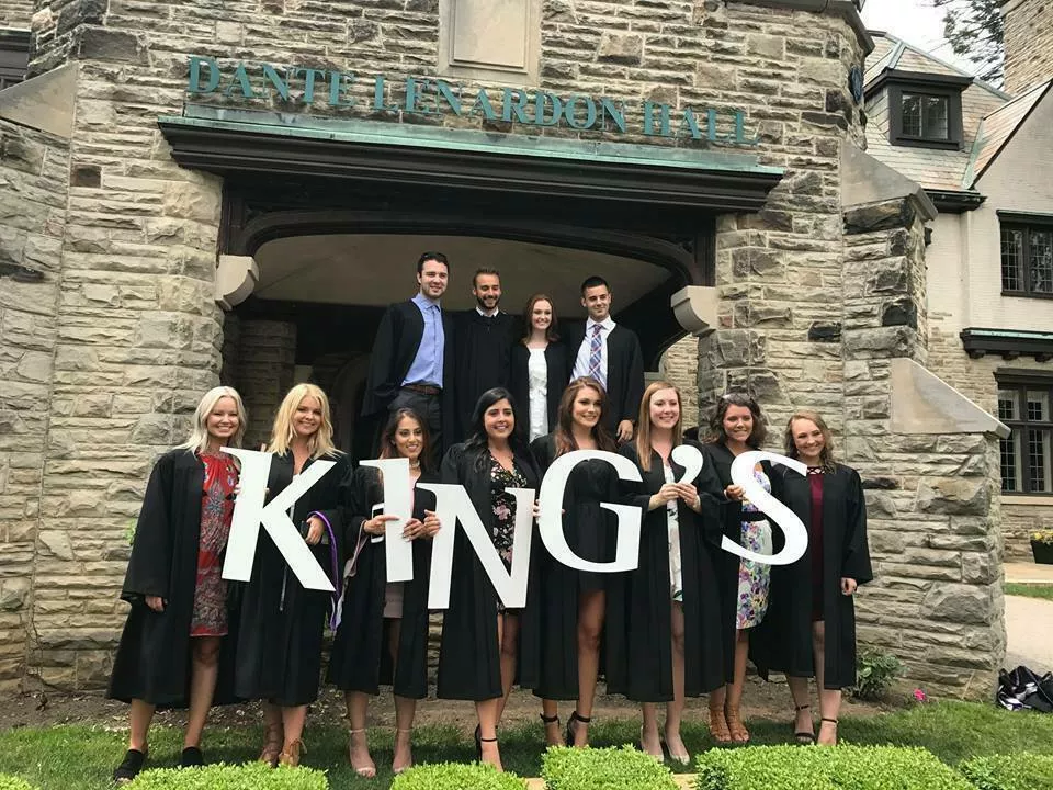 A collection of graduates, some holding large cardboard letters spelling "King's", stand in front of a stone building on King's University College Campus.
