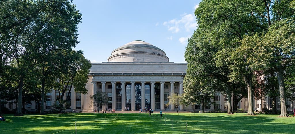 A grey stone building with columns and an impressive central dome is framed by tall stands of deciduous trees and a very green lawn on the MIT campus.