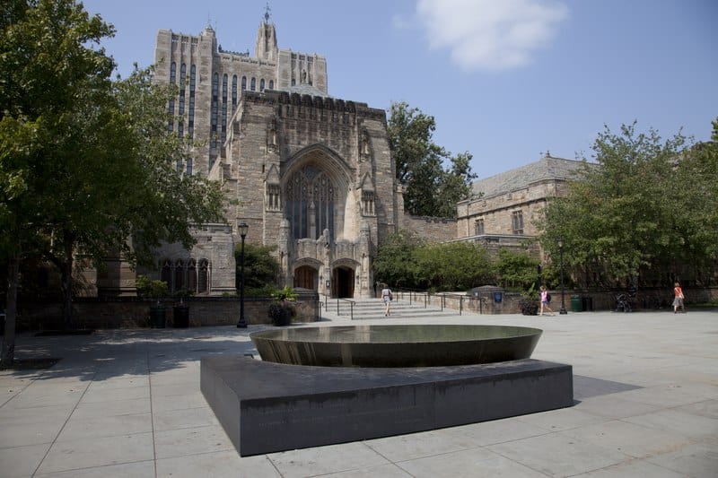 An impressive stone building stands behind a fountain and a large paved square on the Yale campus.