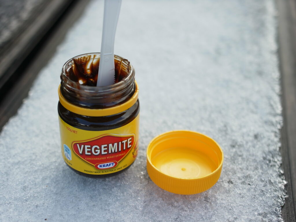 Australian food - an open jar of Vegemite with a plastic knife stuck in it sits next to its lid on an icy surface.