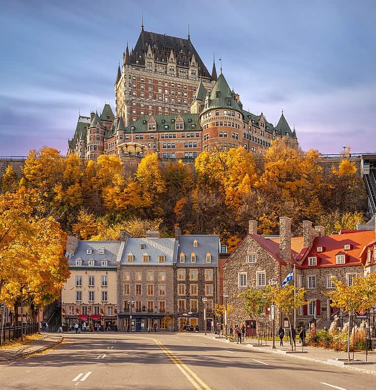 A fall view of Quebec City; a two lane road curving along a city street with three-storey brick historical buildings, a hill with trees in fall foliage, and an imposing castle-like hotel atop the hill.