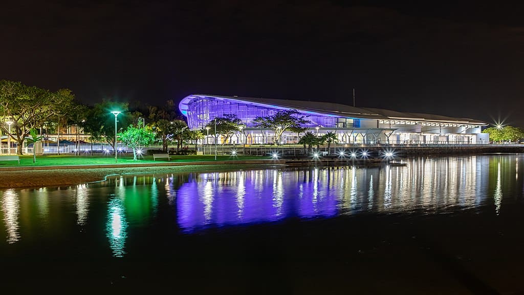 A night scene on a city waterfront with a lit walkway and a swooping building lit with colourful lights