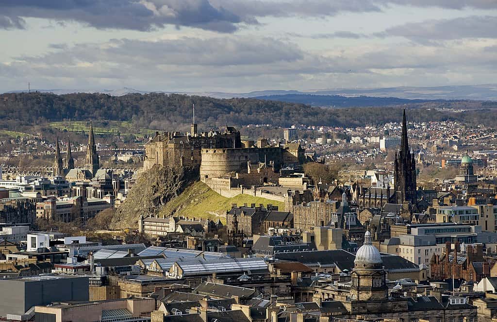 Edinburgh Castle surrounded by the city (glass and steel trainyards, brick cathedrals and government buildings, expanding out to smaller communities with homes. A large forest and tall hills are in the background.