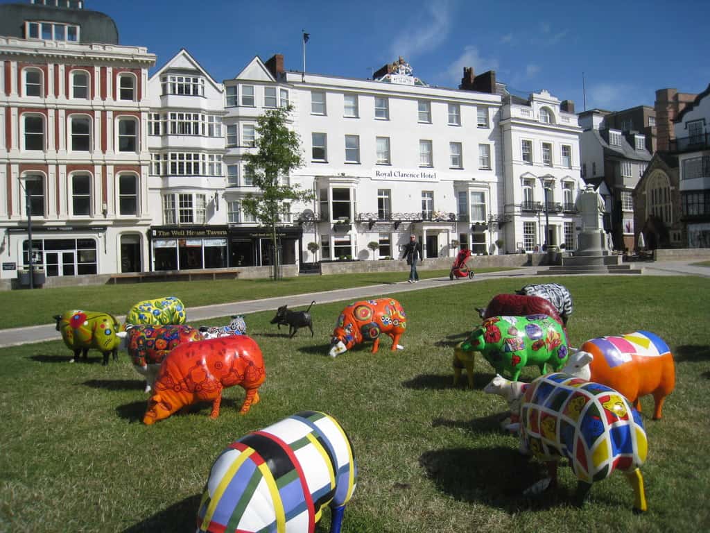 Exeter's Cathedral Close, a lawn surrounded by hotels and businesses (tall, white and brick, multi-storey) with a whimsical flock of brightly painted metal sheep sculptures on the lawn.