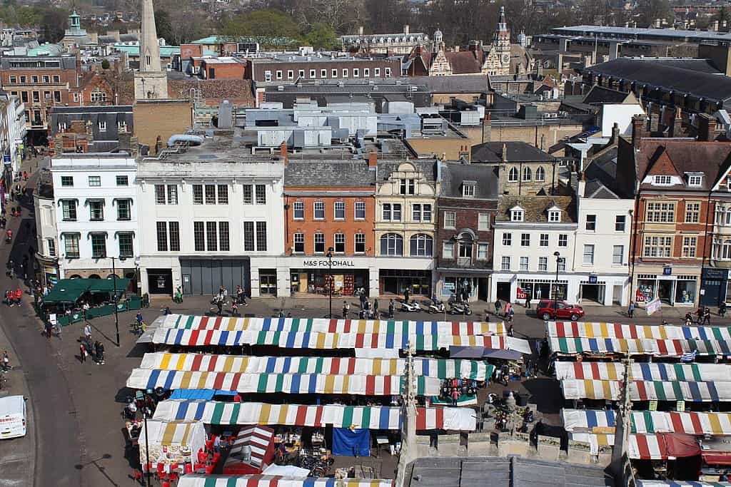 A cityscape of Cambridge (UK): brightly coloured market stalls in the foreground, next to multi-storey brick buildings of different colours. City blocks and clock towers are visible in the background.