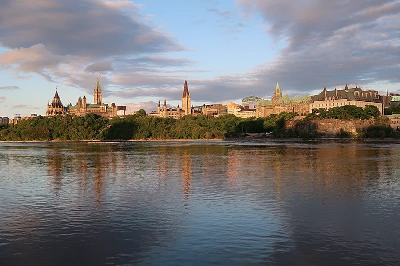 A riverside view of the Ottawa Parliament buildings (sandstone and a green copper roof) under a blue sky with grey clouds.