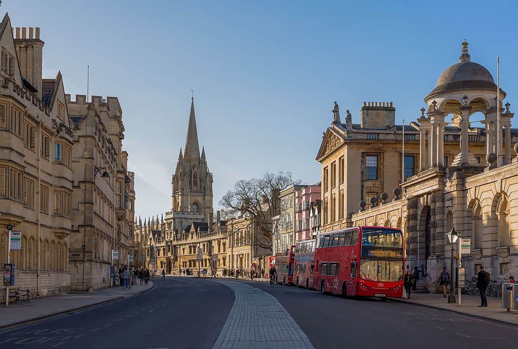 Oxford, UK (a city scene, with parked red tour buses outside an impressive stone building (Queen's College). A cathedral spire towers over the buildings further down the street. 