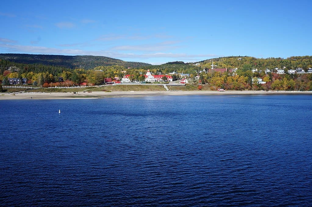 A large lake with blue waters and a blue sky above. A town is nestled at the water's edge. (Saguenay, QC)