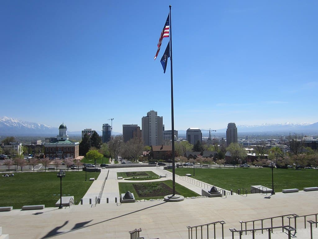 A city scene: taken from the stone steps of a large building with an American flag flying from a flagpole. A number of tall buildings are in the middleground, with the city (Salt Lake City) stretching out behind and to the sides. The city is ringed by snow-capped mountains.