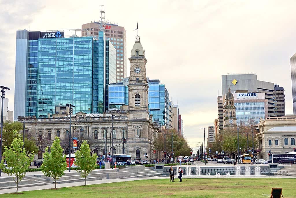 Victoria Square, in Adelaide, AUS is a mostly green lawn with people walking around it and fountains. Early 20th century stone buildings in the middleground; office towers line either side of a busy street.