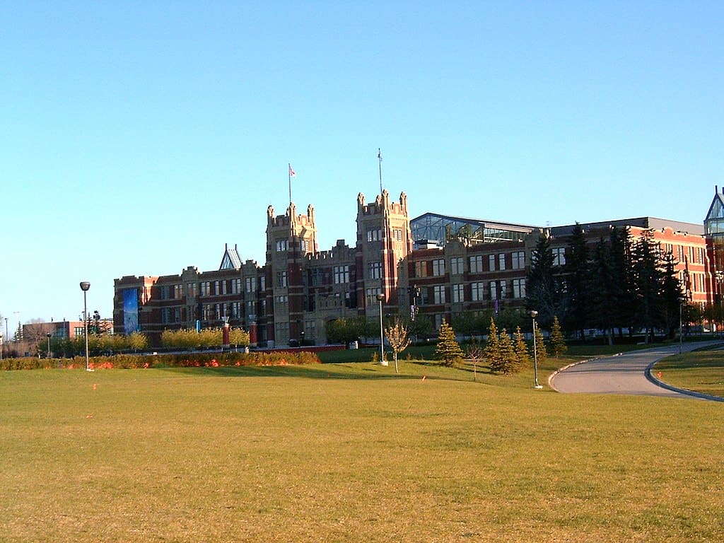 A classic brick building with twin towers at its centre rests at the back of a large lawn, under a blue sky. (SAIT Campus, Alberta)