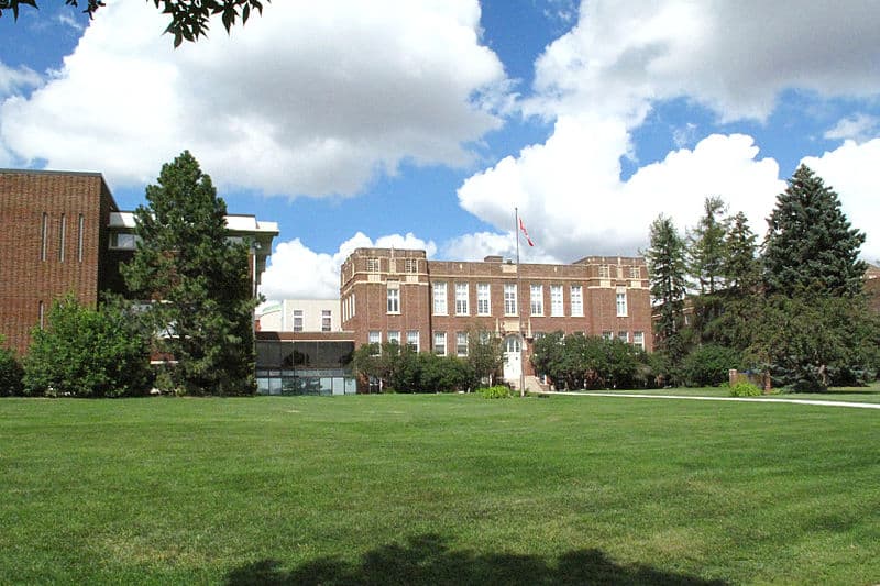 A two-storey brick building sits on a wide green lawn, framed by tall coniferous trees.