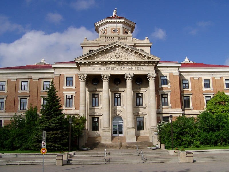 A brick building with Corinthian (ornate) columns flanking the main stairs and door under a blue sky (U of M Admin Building)