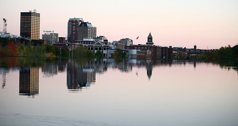 A city skyline with tall and medium-height buildings is contrasted by a river and broad sky at sunrise.