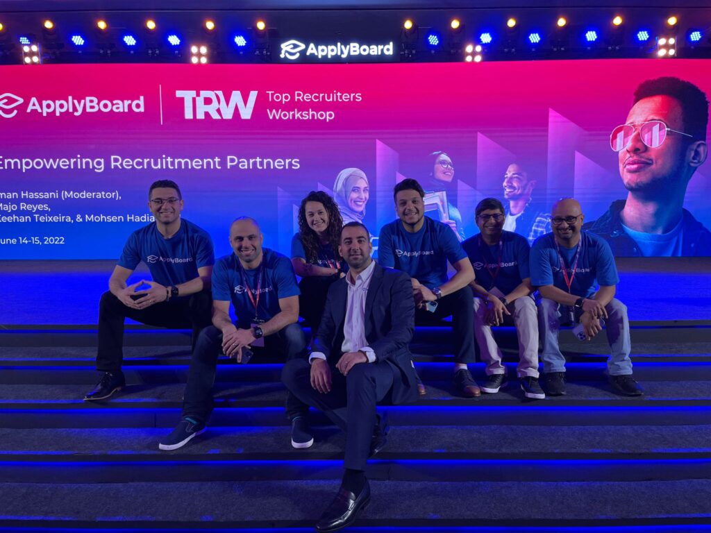 Several members of the ApplyBoard leadership team sitting on blue steps in front of a large TRW panel sign at TRW 2022.
