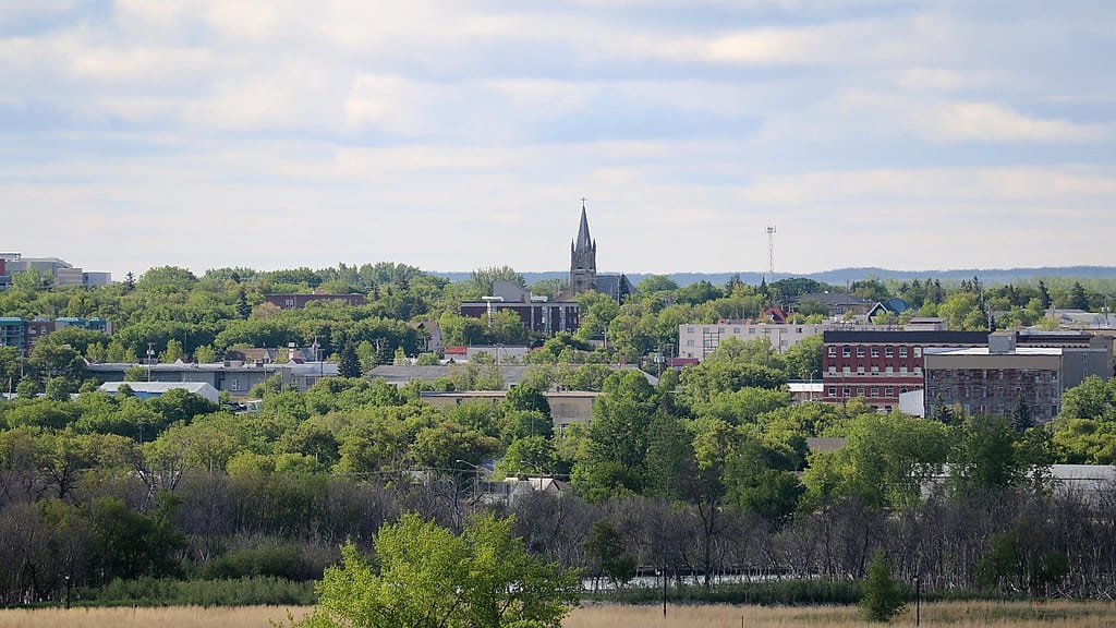 City buildings rise out of a lush green boreal forest; a church's spire is visible on the horizon. (Brandon MB)