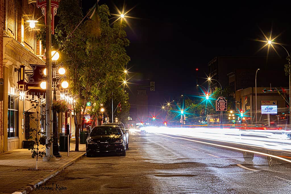 A downtown city street with storefronts, a sidewalk, and multiple exposures blurring oncoming traffic headlights