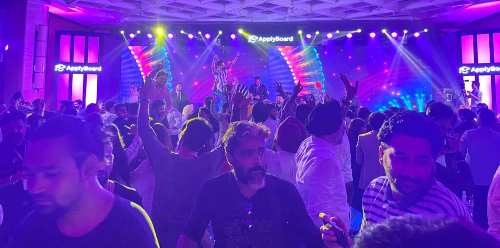 A photograph from TRW 2022 of a hotel room filled with people dining, laughing, and dancing, bathed in purple and blue light, with a band performing on stage in the background.