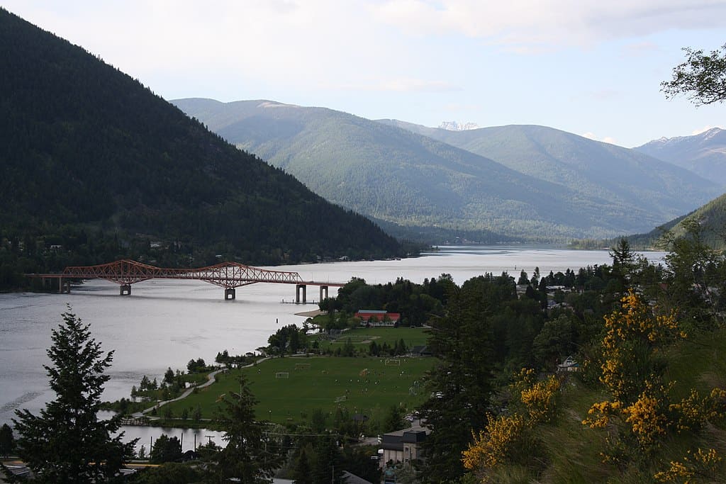 A river valley with mountains on one side and a green field on the other, spanned by a bright orange metal bridge (nelson, british columbia)