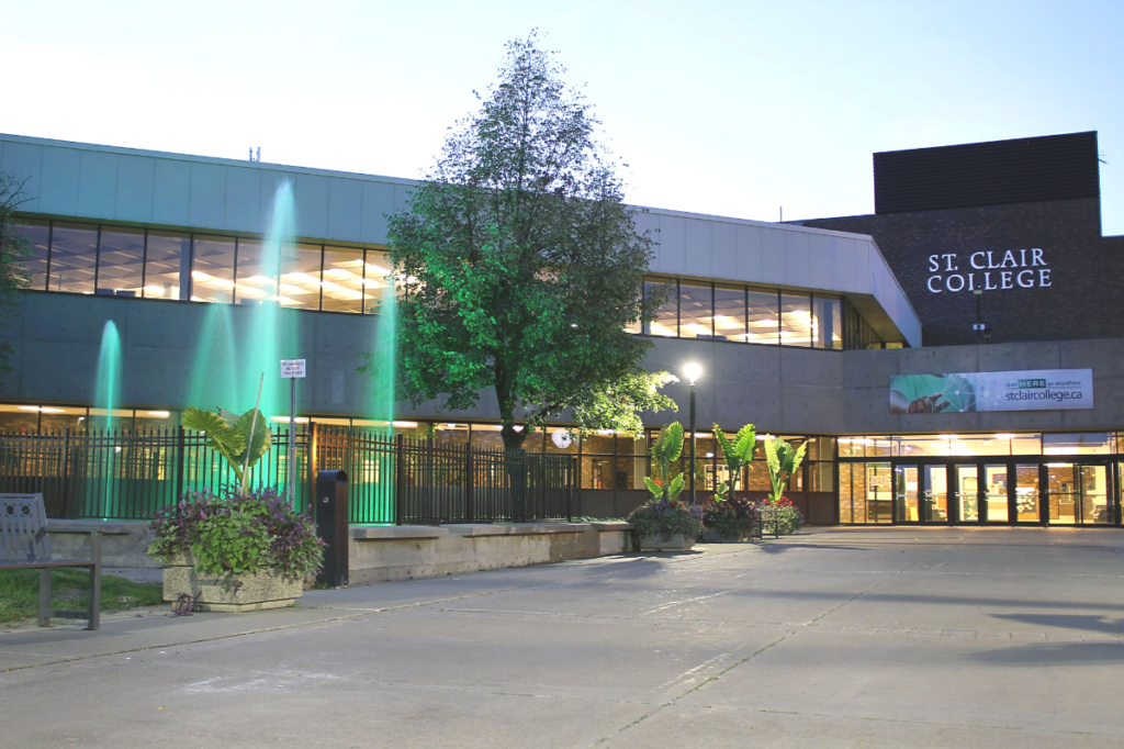An outdoor photo of the St. Clair College main building entrance at dusk, with lit windows, surrounded by greenery, and featuring green backlit water fountains.