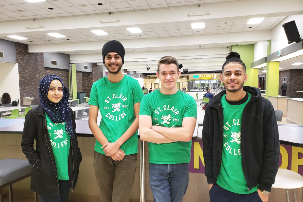 Four St. Clair College students, each proudly wearing a green St. Clair College t-shirt, smiling and standing side-by-side in the College's indoor cafeteria