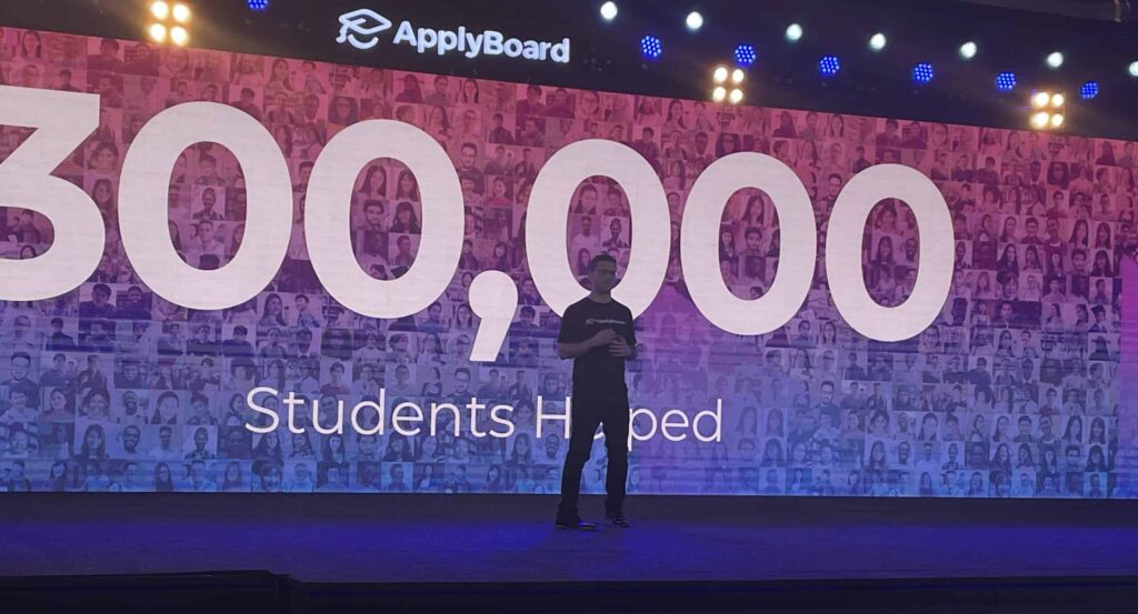 Martin Basiri on stage at TRW 2022, with the words "300,000 Students Helped" displayed behind him over a photo collage of students helped by ApplyBoard.