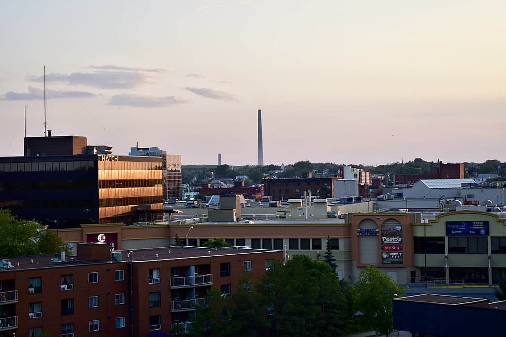 A cityscape at twilight, with low- and medium-height brick and concrete buildings stretching to the horizon, with a mall at the foreground. (Sudbury ON)