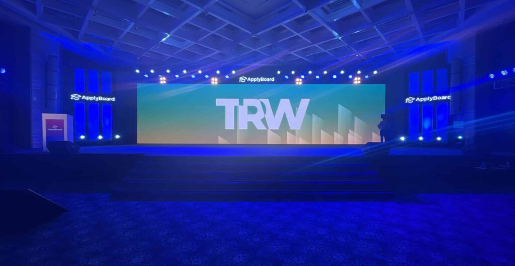 A photo of the empty hotel conference room at TRW 2022, bathed in blue light, with "TRW" displayed against a colourful backdrop on the main event stage.