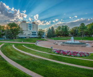 A photograph of Colorado State University-Pueblo with clouds surrounding a building and a fountain with green grass in the centre.