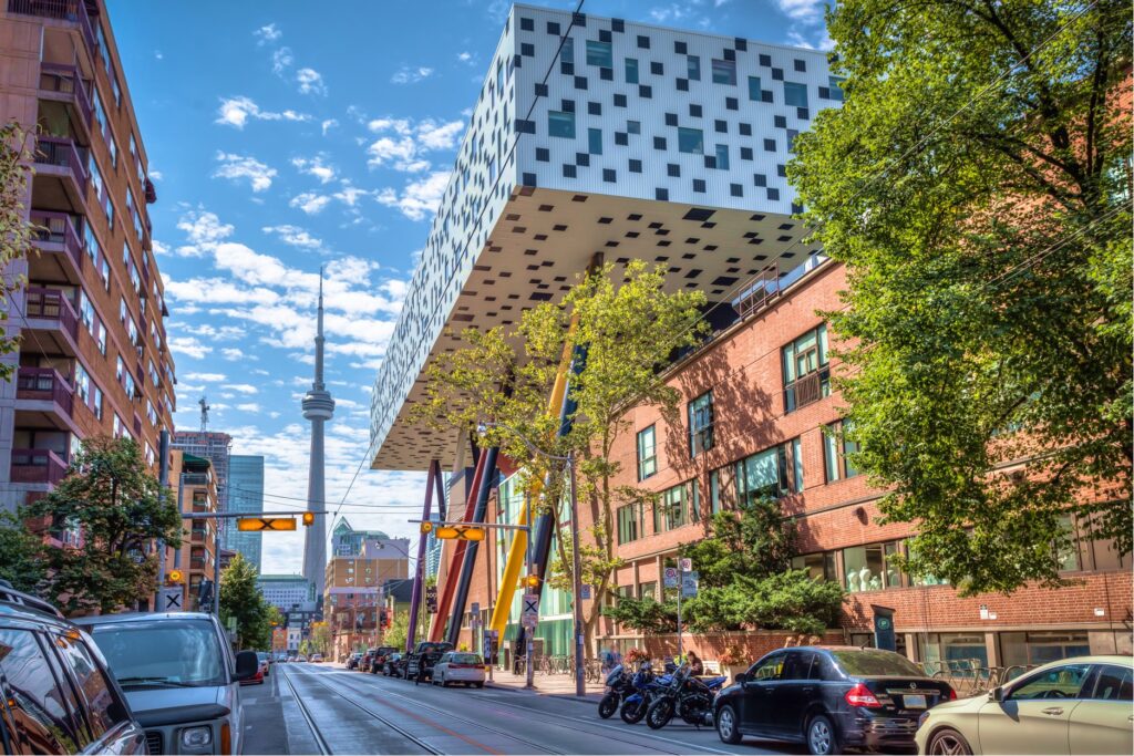 A view of OCAD University and the CN Tower in Toronto (bright cityscape; needle tower visible at the end of a street against a blue sky; OCAD in the foreground - a silver, suspended building with lots of square windows.)
