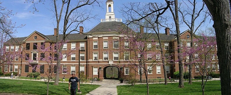 A long, three-storey red brick building sits on a green lawn. There are plum trees coming into blossom in front of the building.