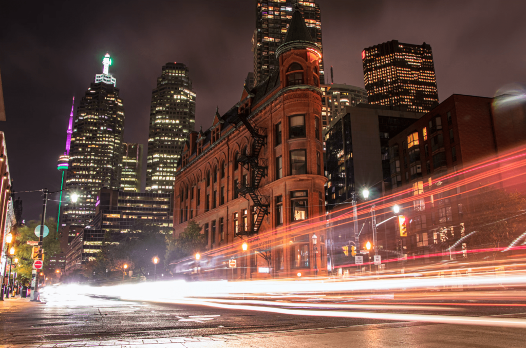 A night scene of Toronto, Canada; the Gooderham Building (red brick flatiron) in the foreground; headlights blur past, the CN Tower shines in the background.