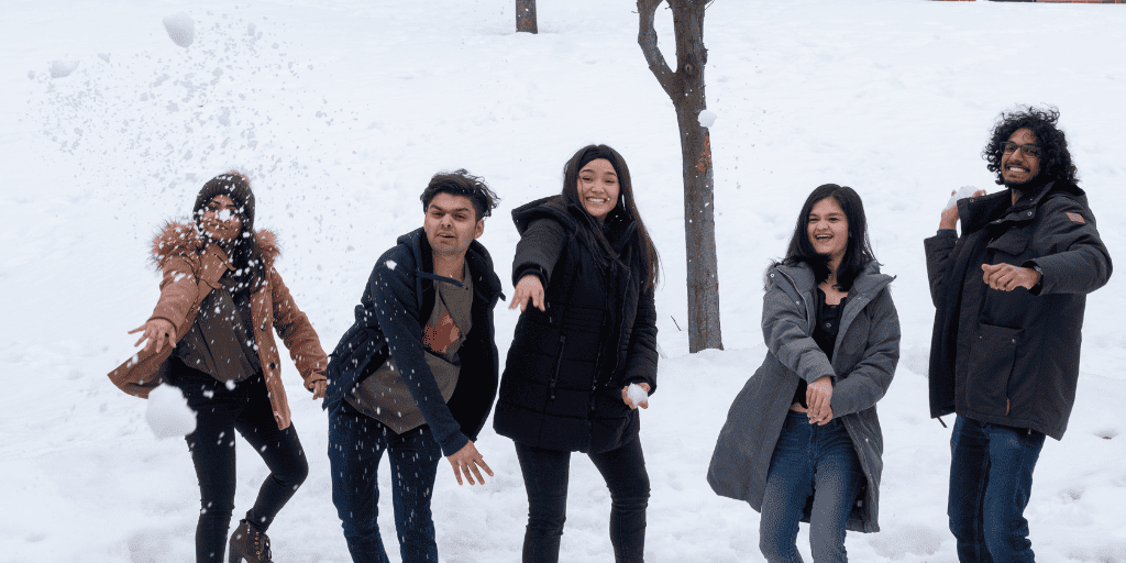Five college students in warm winter clothing stand in a line in a snowy field; one of them has just thrown a snowball.