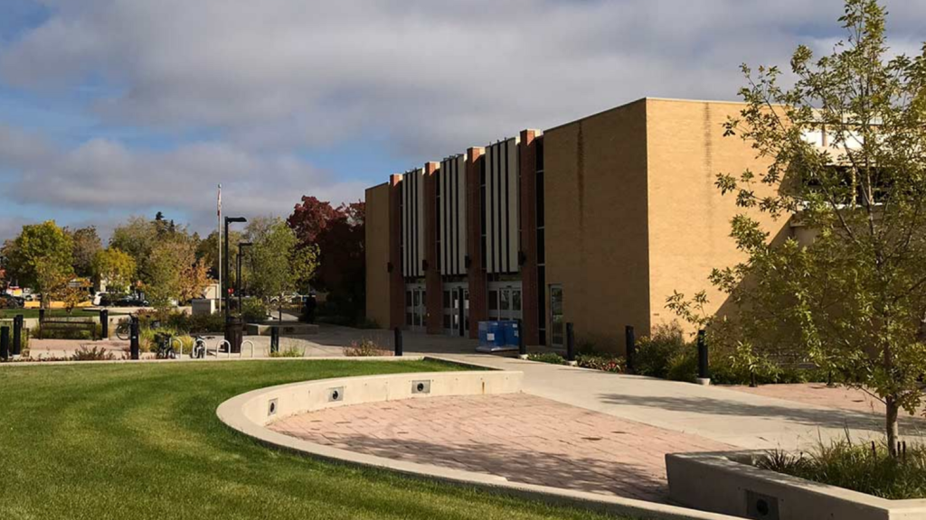 A multistorey blond brick building sits at the edge of a curved lane, edged by a green grass lawn (Saskatchewan Polytechnic campus)
