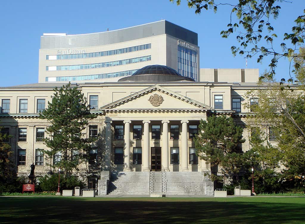 A three-storey university building with a columnaded entry sits on a green lawn framed by trees. A more modern university building on the UOttawa campus rises behind it.